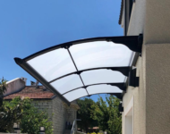 Curved transparent awning attached to a beige house, shielding a doorway outdoors.