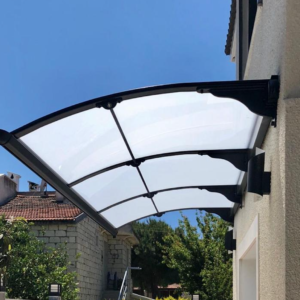 Curved transparent awning attached to a beige house, shielding a doorway outdoors.