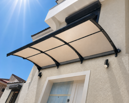 Beige house front with a curved translucent canopy over a white door framed by a black metal frame; clear blue sky and sun glare above.