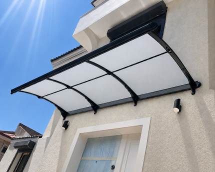 Curved translucent canopy over a front door on a beige stucco wall, with black metal supports and a bright blue sky above.