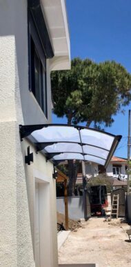 Exterior of a light stucco building with a curved translucent canopy over the entry, supported by black brackets; construction area visible in the background.