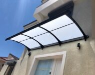 Curved translucent canopy with a black metal frame mounted above a white-framed window on a beige house, against a blue sky.