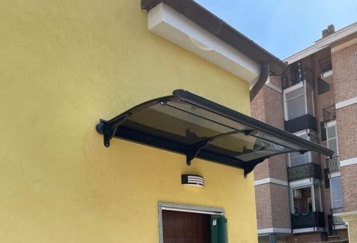 Black glass canopy extending over a doorway on a yellow stucco wall, with a small outdoor light fixture above and a brick building in the background.
