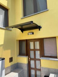 Yellow building entrance with a brown framed door under a black metal canopy and a small wall light above, gray stone base and window sills nearby.