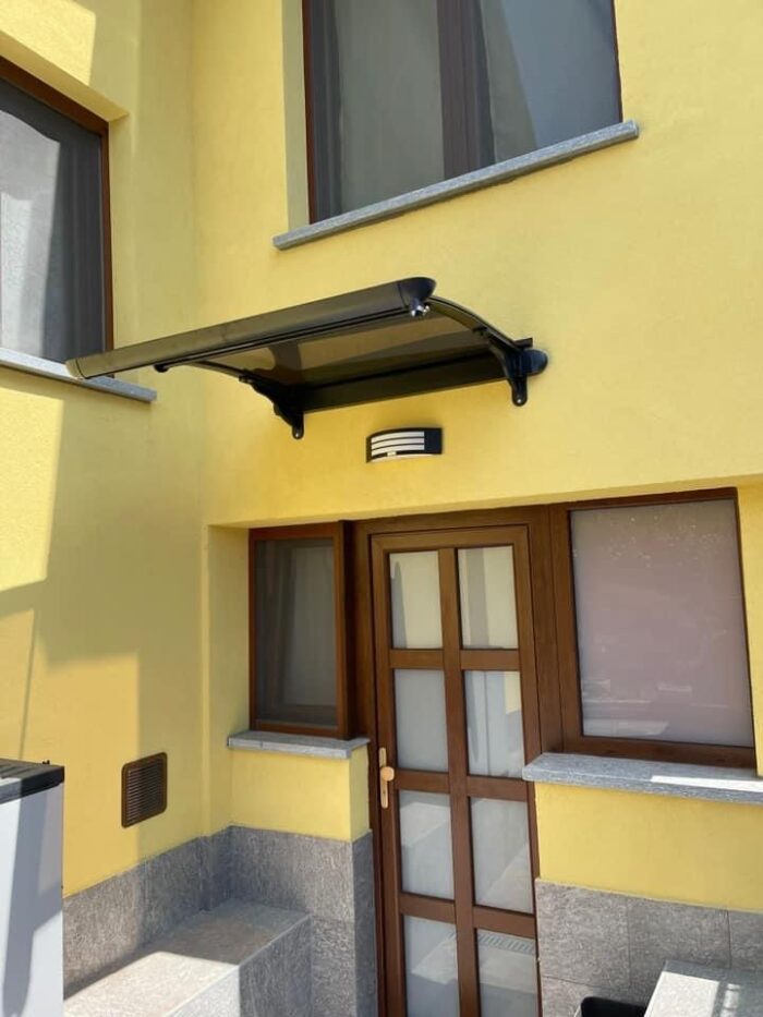 Yellow building entrance with a brown framed door under a black metal canopy and a small wall light above, gray stone base and window sills nearby.
