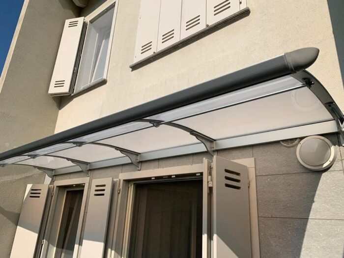 Exterior of a house with a curved translucent awning over a doorway and white window shutters above.