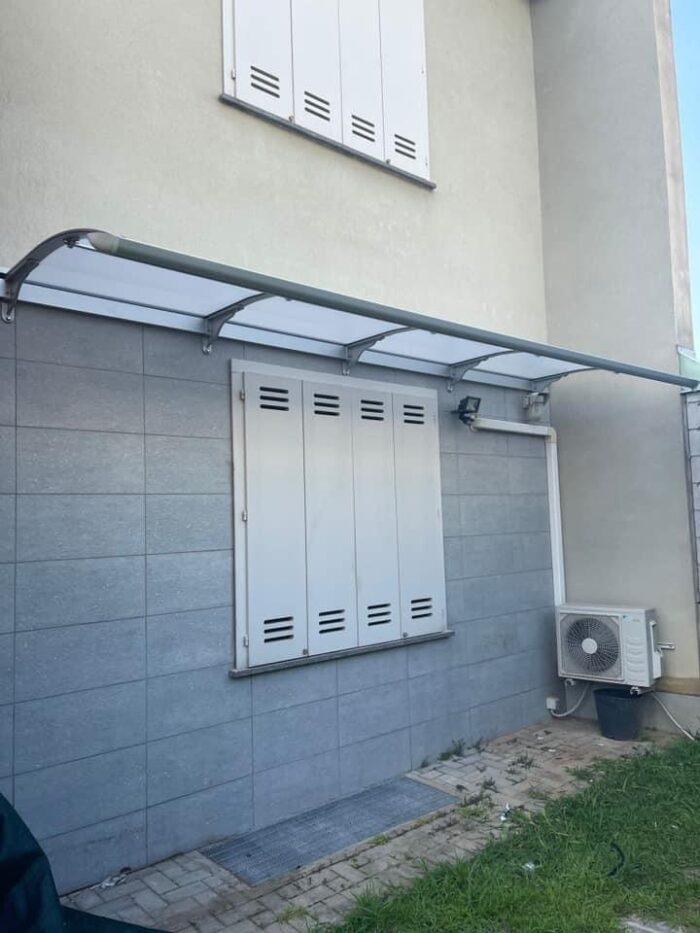 Exterior wall with four-panel white vented shutters, a glass canopy, and an outdoor air conditioner unit on the right side.