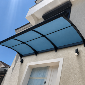 Curved blue-tinted glass canopy with black metal frame above a white door on a beige stucco wall, under a clear blue sky.