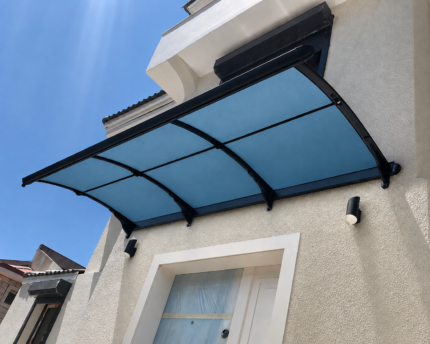 Curved blue-tinted glass canopy with black metal frame above a white door on a beige stucco wall, under a clear blue sky.